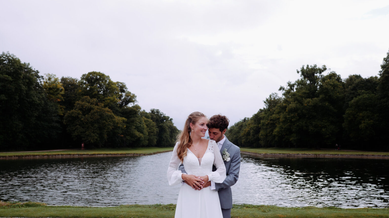 Pareja internacional de recién casados posando frente a un río en un parque, con un entorno natural y sereno como fondo. En los parques del Palacio de Nymphenburg, Munich